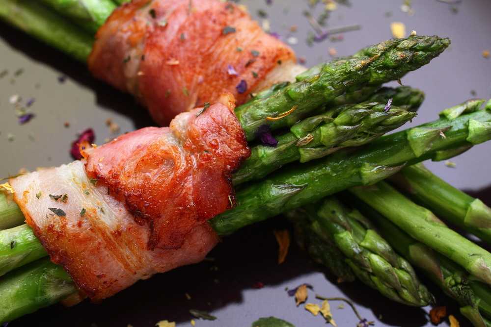 Asparagus stems wrapped with crispy bacon on oily plate topped with ground black pepper