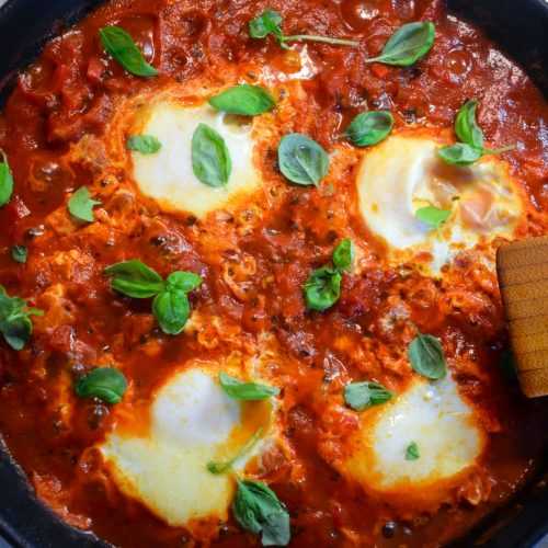 Four Cooked eggs inside a red sauce topped with basil leaves in a black pan