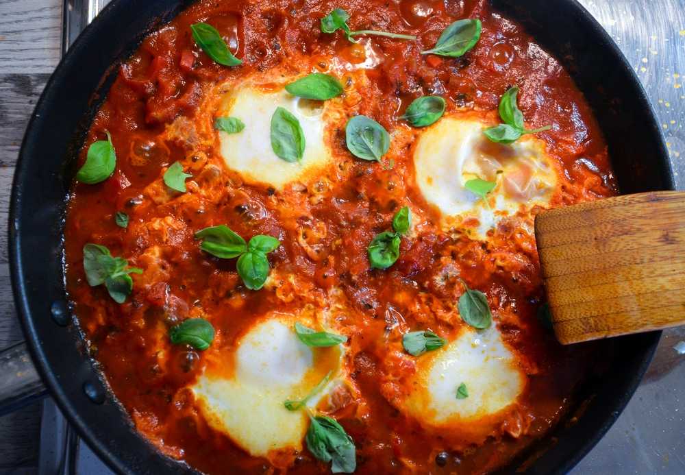 Four Cooked eggs inside a red sauce topped with basil leaves in a black pan 