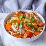 Udon noodles with carrot, red bell pepper, chicken and onion topped with chopped parsley in white bowl