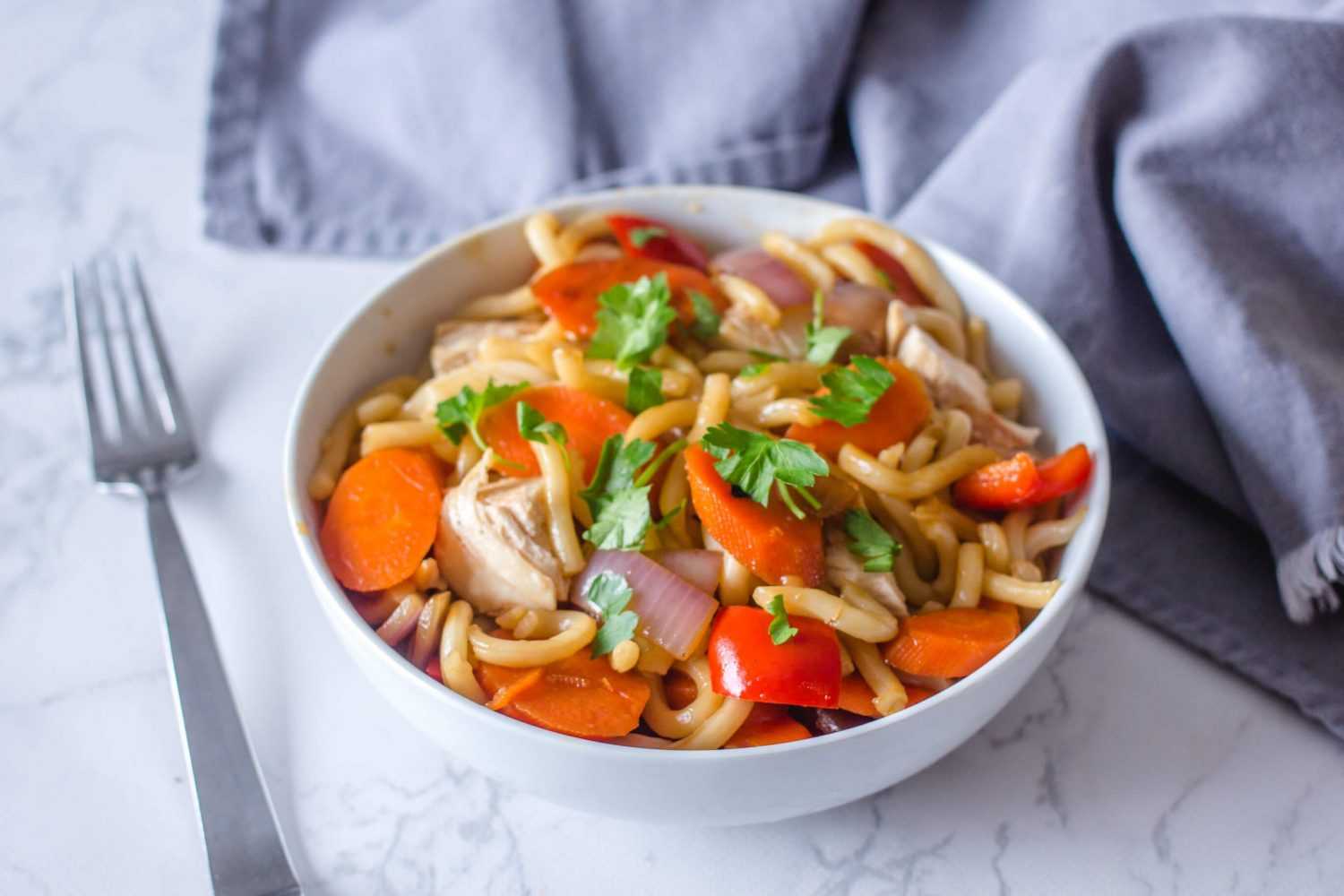 Udon noodles with carrot, red bell pepper, chicken and onion topped with chopped parsley in white bowl