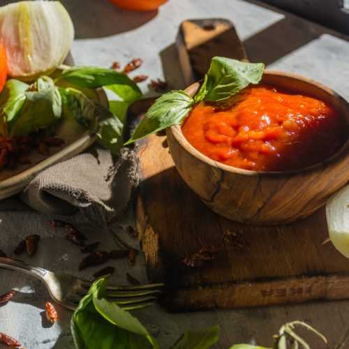 Wooden bowl filled with Arrabiata sauce with basil, tomatoes and onion on side