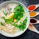 Clear soup of chicken and rice noodles topped with mint, coriander and beansprouts with spoon with sauces