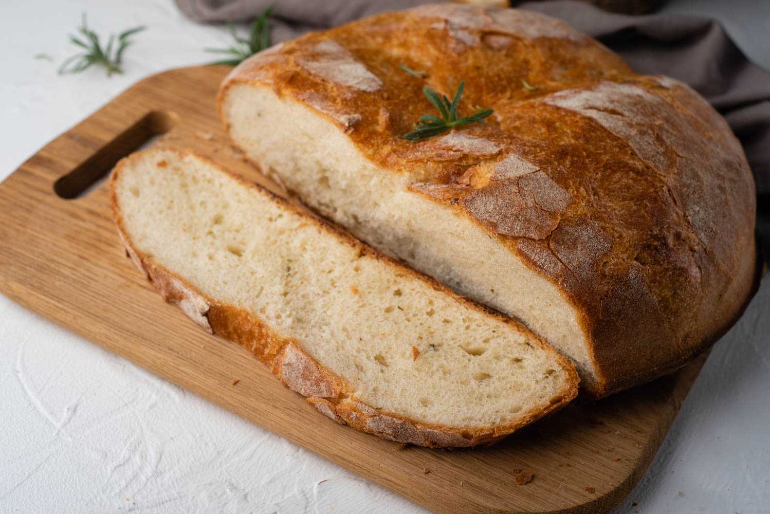 Whole rosemary bread with one slice cut out on cutter board topped with rosemary