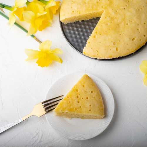 Slice of vanilla cake on a small white plate near a whole cake with yellow flowers on side