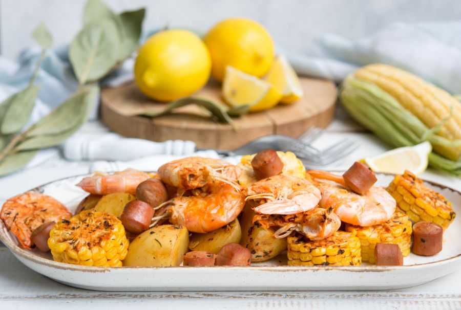 Mix of corn ears, shrimp, potato cubes and slices sausage in a white plate with lemon and corn on side