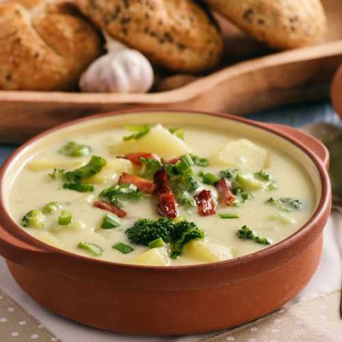 Creamy soup in brown bowl filled with chopped potatoes, broccoli florets, bacon strips and chopped scallion