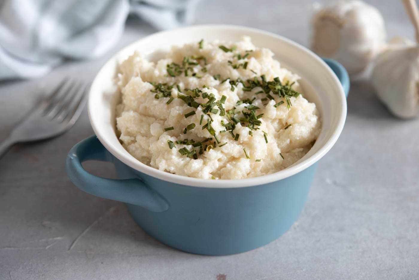 Cauliflower Mush served in a blue bowl topped with chopped chives and whole garlic on side