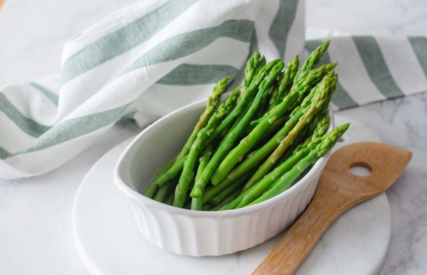 Asparagus spears in white bowl next to a wooden spoon