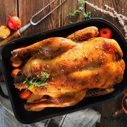 Whole duck in a black pan on wooden table with carrots, tomatoes, rosemary and spices