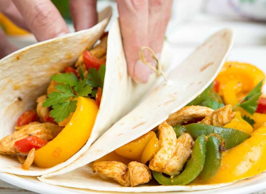 Hand holding tortilla filled with chicken strips, green and yellow bell pepper and parsley with another tortilla on side