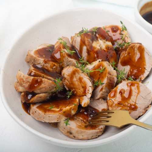 Pork medallions glazed with soy sauce in a bowl with metal fork and sage leaves