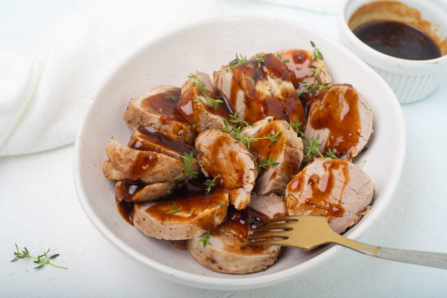 Pork medallions glazed with soy sauce in a bowl with metal fork and sage leaves