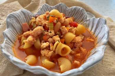 Macaroni in red soup mixed with ground turkey, chopped carrot and celery and sausage pieces in white bowl