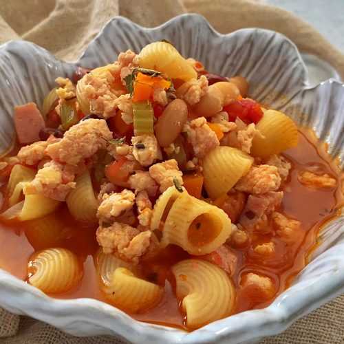 Macaroni in red soup mixed with ground turkey, chopped carrot and celery and sausage pieces in white bowl