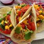 Four open tacos filled with shredded chicken, cherry tomatoes, mango and avocado cubes and parsley on white plate