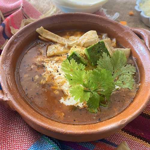 Brown soup filled with shredded chicken, tortilla strips and carrot cubes topped with parsley in brown bowl