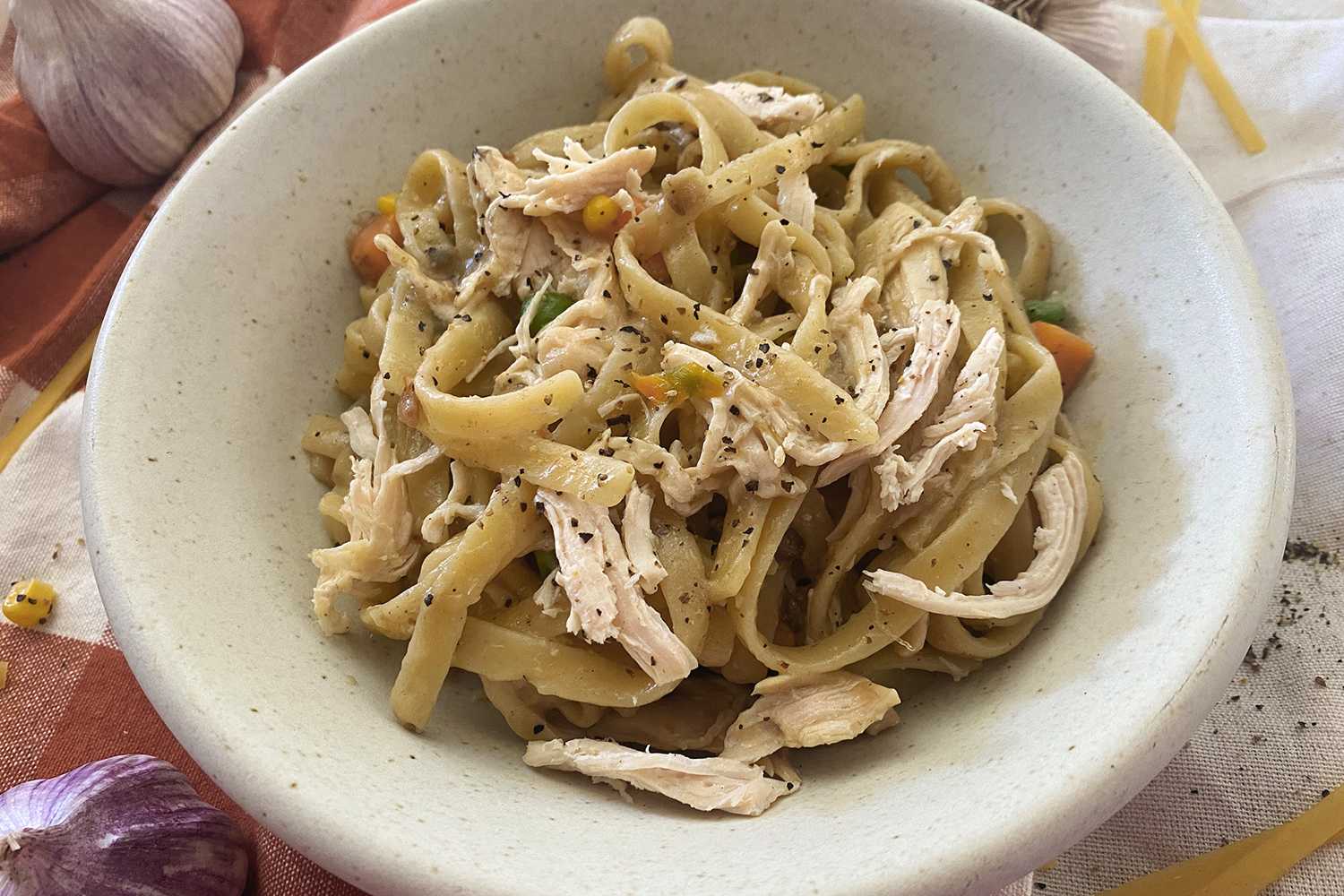 Fettuccine pasta with chicken strips, corn, peas and chopped carrot in white bowl.