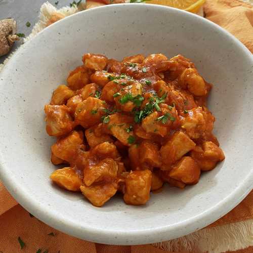 Chicken cubes in an orange sauce topped with chopped parsley in a white bowl with orange slices on side