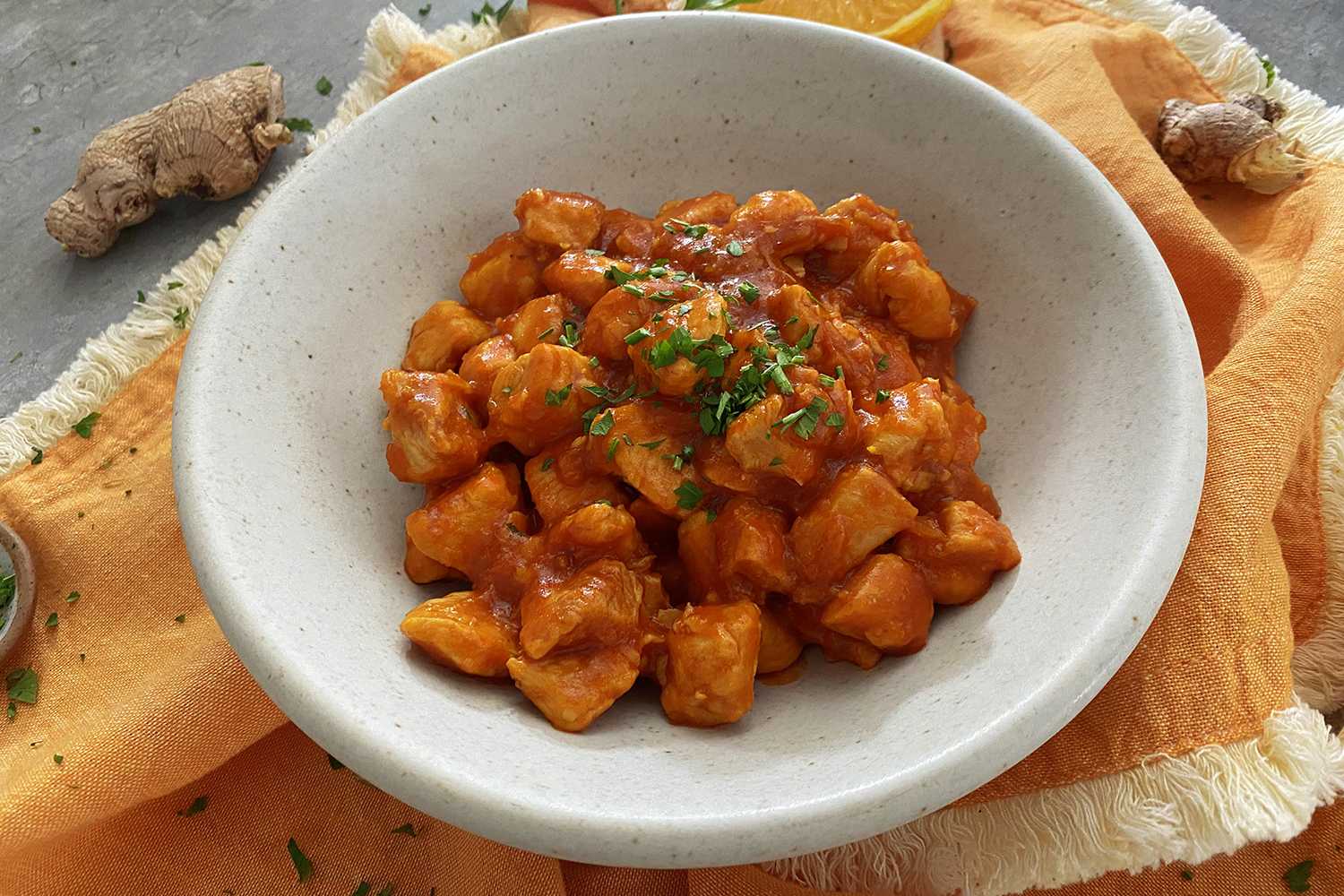 Chicken cubes in orange sauce topped with chopped parsley in a white bowl with ginger on side
