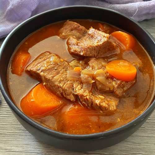 Clear soup with beef steak, carrot cubes and chopped onion in a black bowl