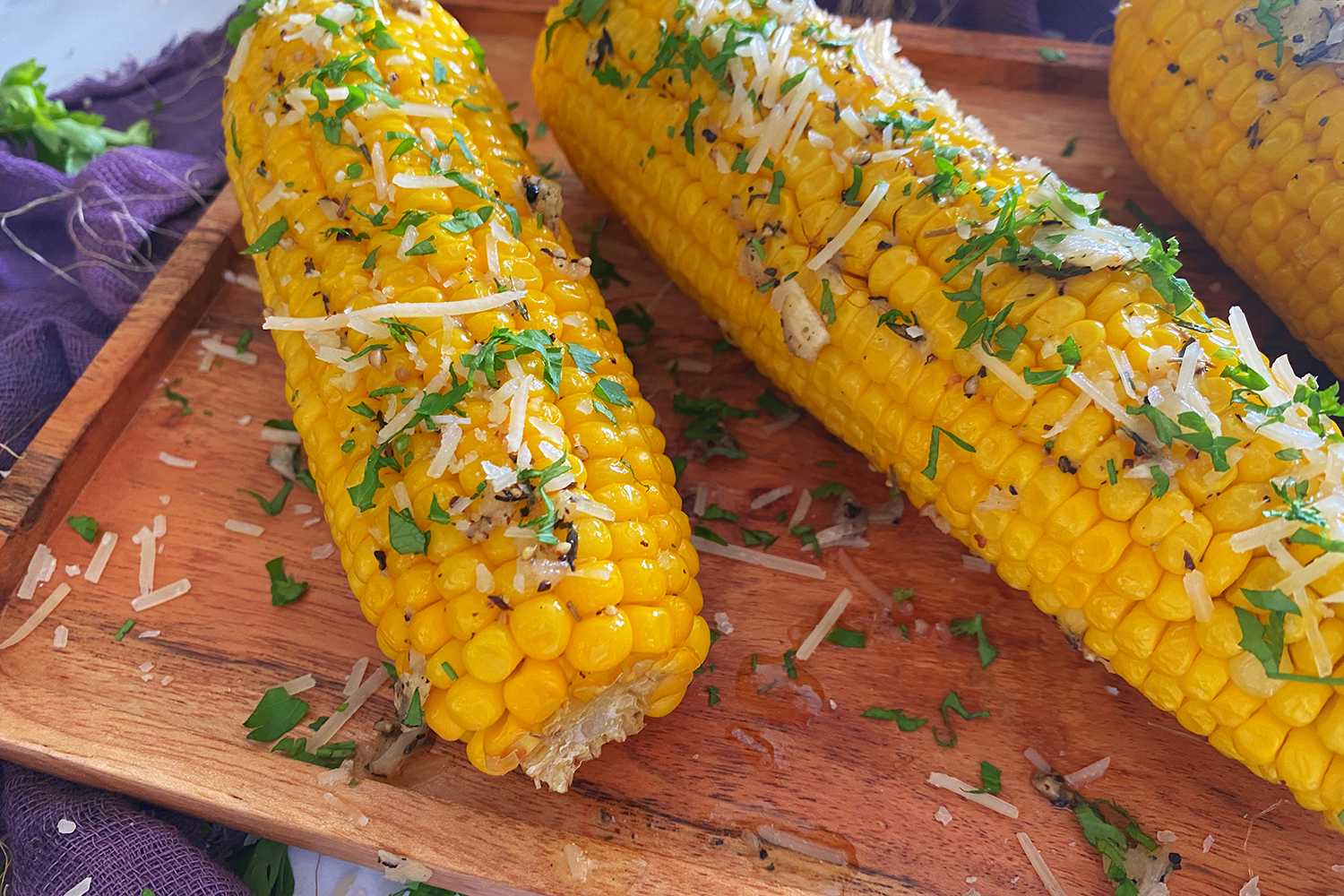 Two ears of corn topped with cheese, salt and chopped parsley on a cutter board
