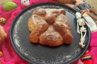 Homemade Pan de Muerto with flour on top on a black plate and plastic skulls on side