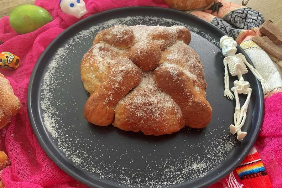 Homemade Pan de Muerto with flour on top on a black plate and plastic skulls on side