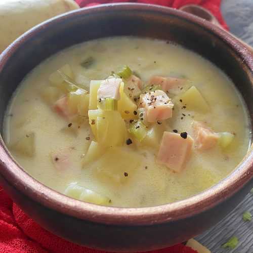 Yellow soup filled with ham cubes, potato cubes, celery and onion in brown bowl