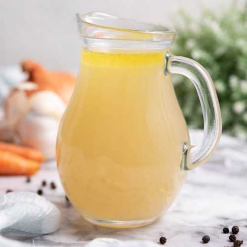 A jug full with homemade chicken stock near a carrots and veggies on a gray table