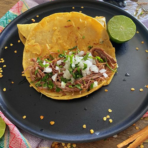 open birria taco filled with beef chunks, chopped onion and parsley on a black plate