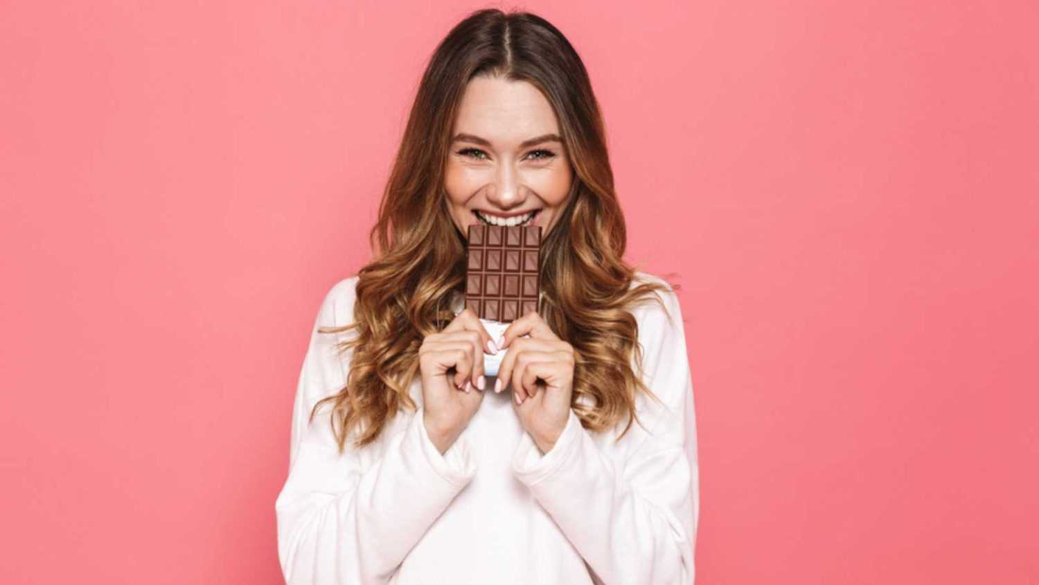 Portrait of a happy young woman biting chocolate bar isolated over pink background