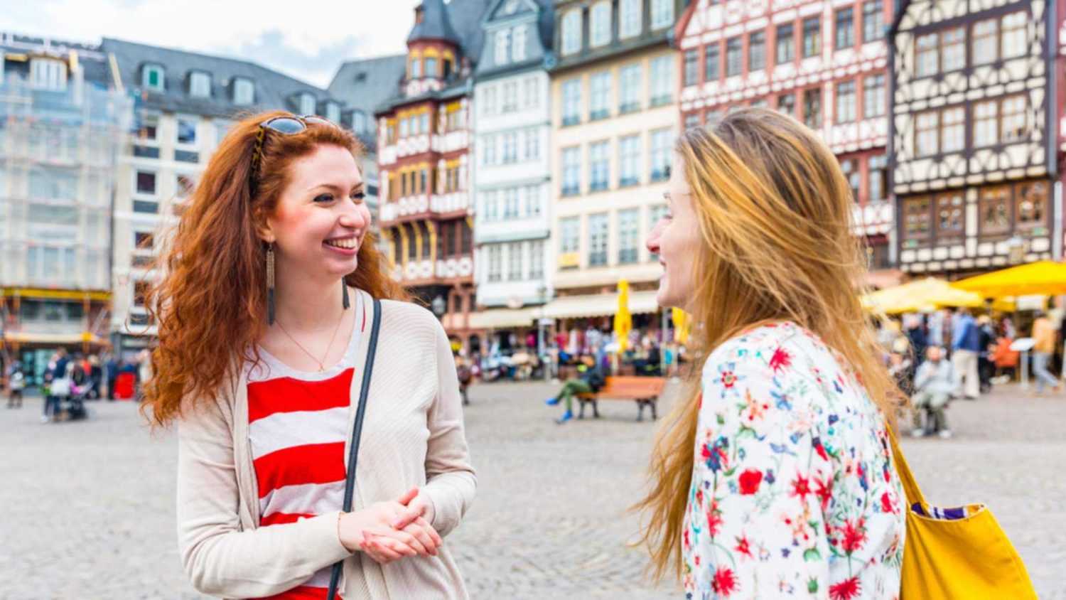 Two beautiful women meeting and having fun in Frankfurt main square. Girls smiling and chatting with houses and people on background. Friendship and travel concepts.