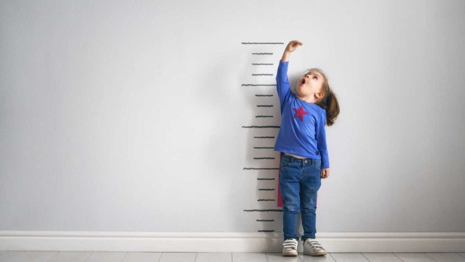 Little child is playing superhero. Kid is measuring the growth on the background of wall. Girl power concept.