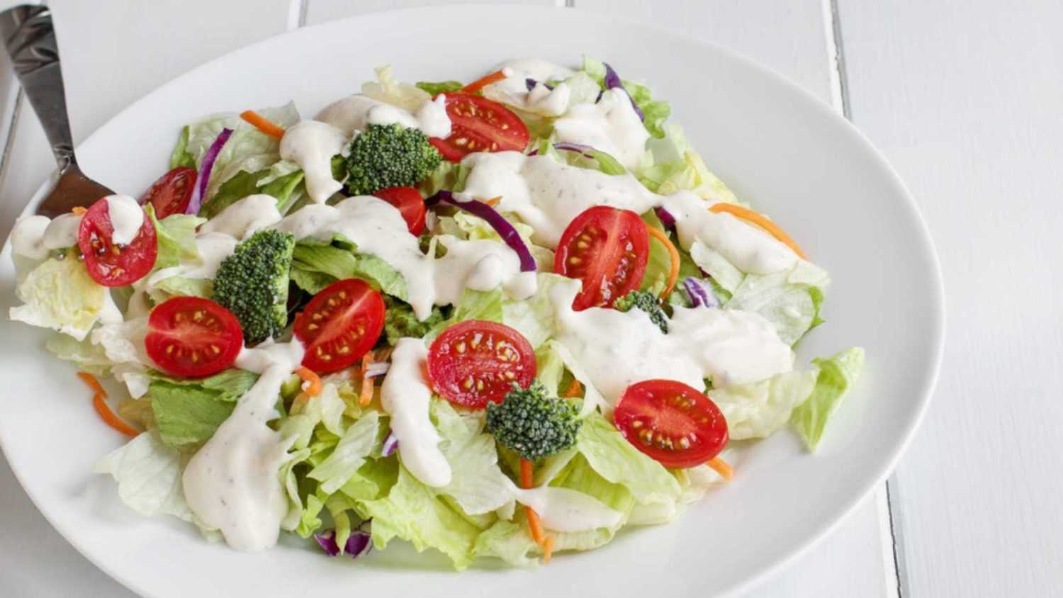 Plate of homemade fresh salad with buttermilk ranch dressing, tomatoes, broccoli, cabbage and carrots served over a white wooden table. House Salad.