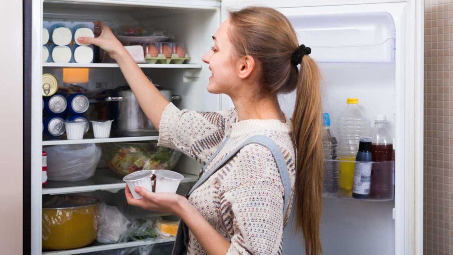 Happy young longhaired woman arranging products on fridge shelves and smiling