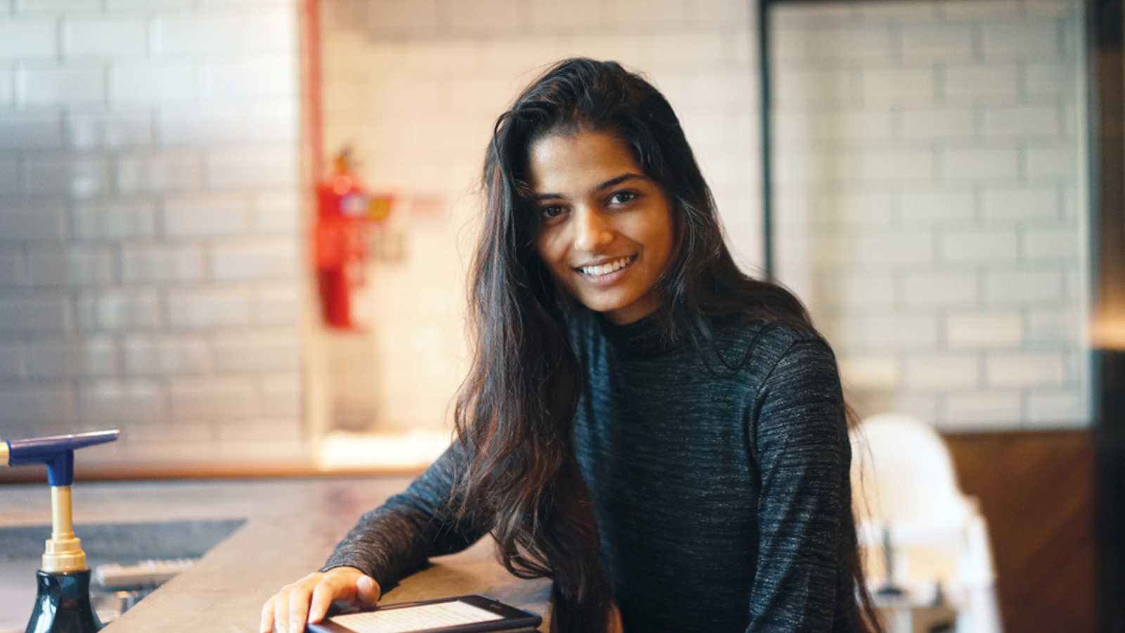 Portrait of a young attractive Indian woman sitting at a table in a cafe, casually and naturally smiling up from her kindle.