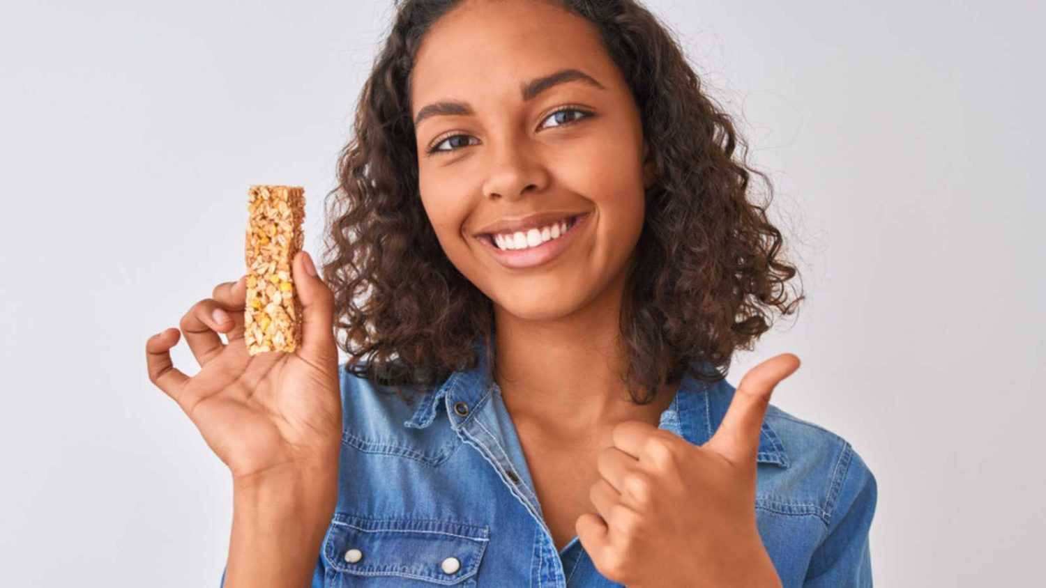 Young brazilian woman eating granola bar standing over isolated white background happy with big smile doing ok sign, thumb up with fingers, excellent sign