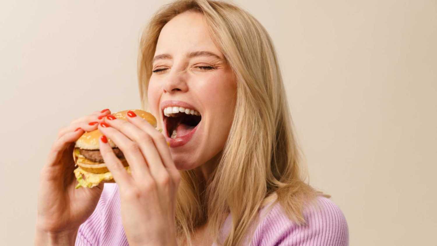 Happy beautiful hungry girl eating hamburger on camera isolated over white background