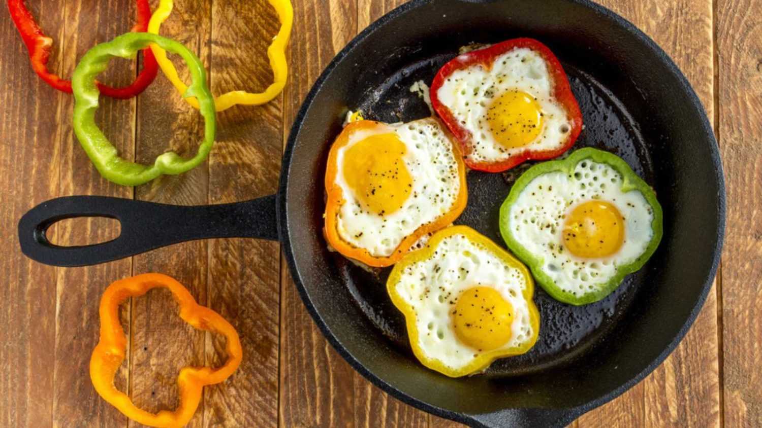 Large cast iron skillet with fried eggs in green, yellow, red and orange bell peppers sitting on wooden table with pepper slices