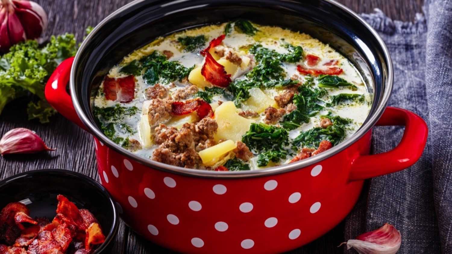 Zuppa Toscana, hearty Tuscan Soup loaded with Italian sausage, kale, fried bacon and potatoes in red pot on dark wooden table, close-up